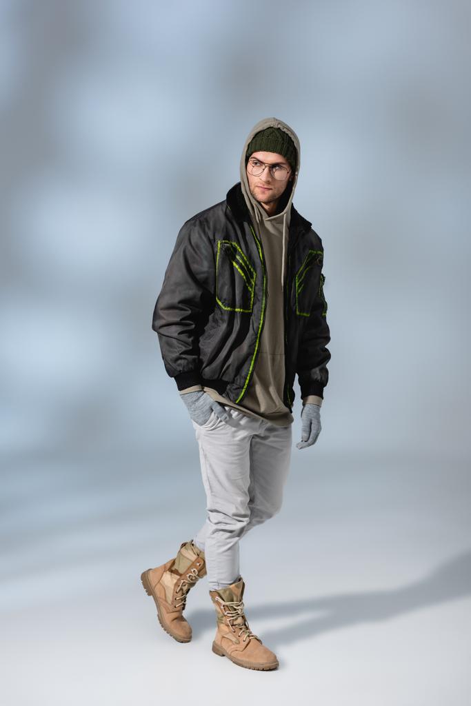 full length of trendy man in hat, hoodie and anorak posing on grey - Photo, Image