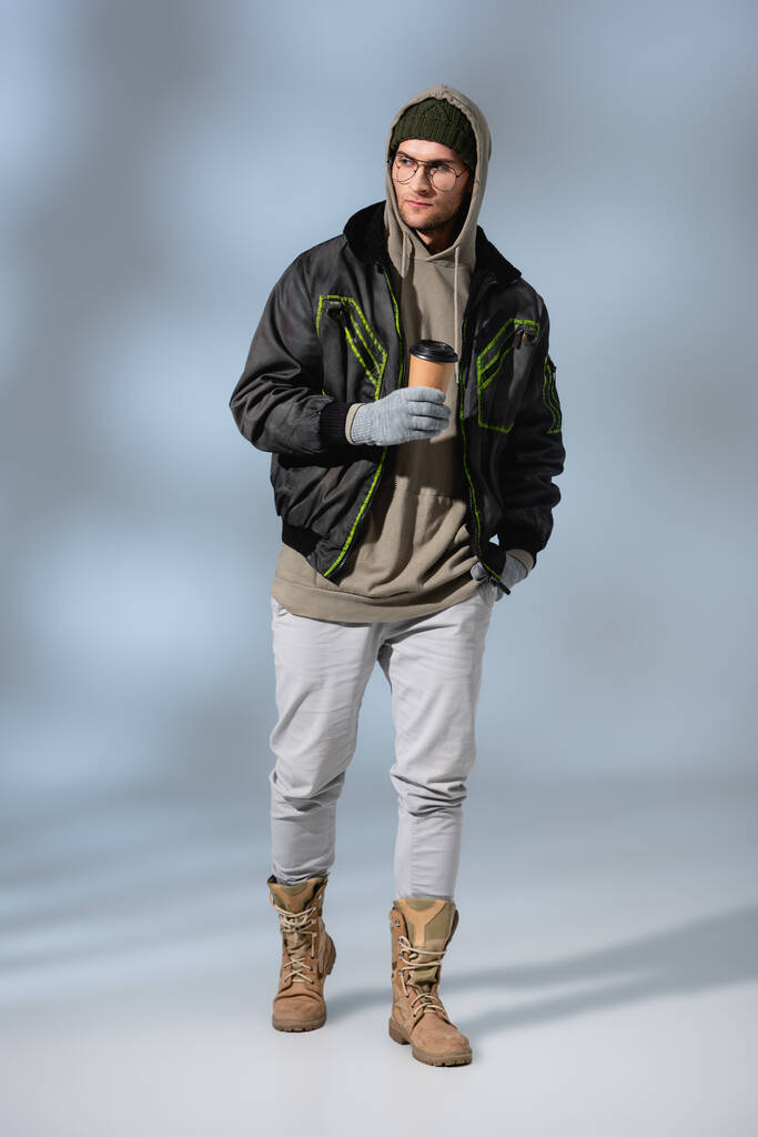 full length of young trendy man in hat, hoodie and anorak ποζάρουν με χάρτινο κύπελλο στο γκρι - Φωτογραφία, εικόνα