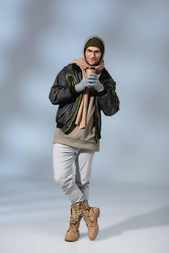 Aufgeregter Mann mit Hut, Kapuzenpulli und Anorak steht mit überkreuzten Beinen und hält Pappbecher auf grau - Foto, Bild