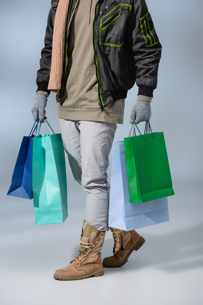 vue partielle de l'homme élégant en tenue d'hiver tenant des sacs à provisions sur gris - Photo, image