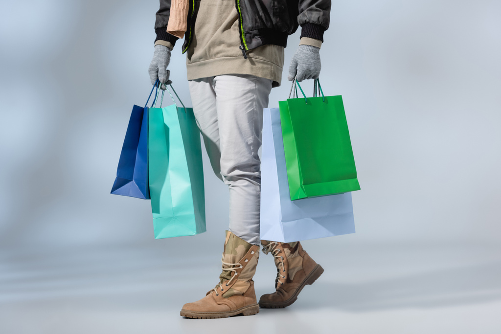 vista recortada de hombre con estilo en traje de invierno sosteniendo bolsas de compras en gris - Foto, Imagen