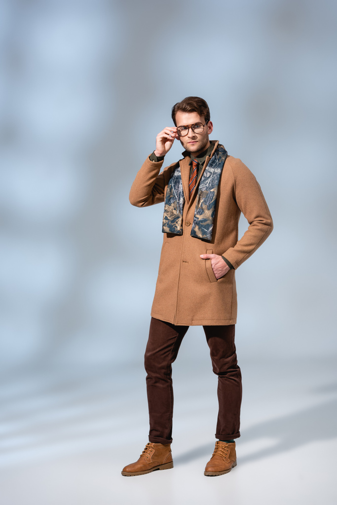 full length of trendy man in winter coat adjusting glasses while standing with hand in pocket on grey - Photo, Image