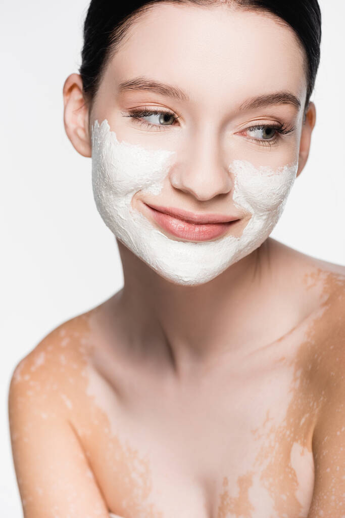sonriente joven hermosa mujer con vitiligo y máscara de arcilla en la cara aislada en blanco - Foto, imagen