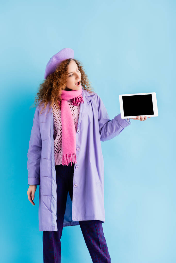 mulher espantada no casaco de inverno, boina e lenço de malha rosa olhando para tablet digital com tela em branco no azul - Foto, Imagem