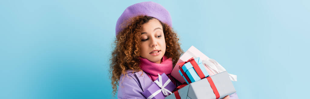 encaracolado jovem mulher em casaco de inverno, boina e lenço de malha rosa segurando monte de presentes de Natal em azul, banner - Foto, Imagem