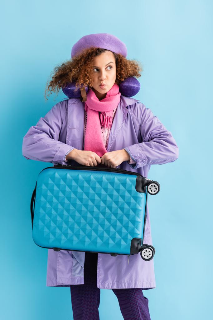 mladá kudrnatá žena v kabátě a šále drží kufr na modré - Fotografie, Obrázek