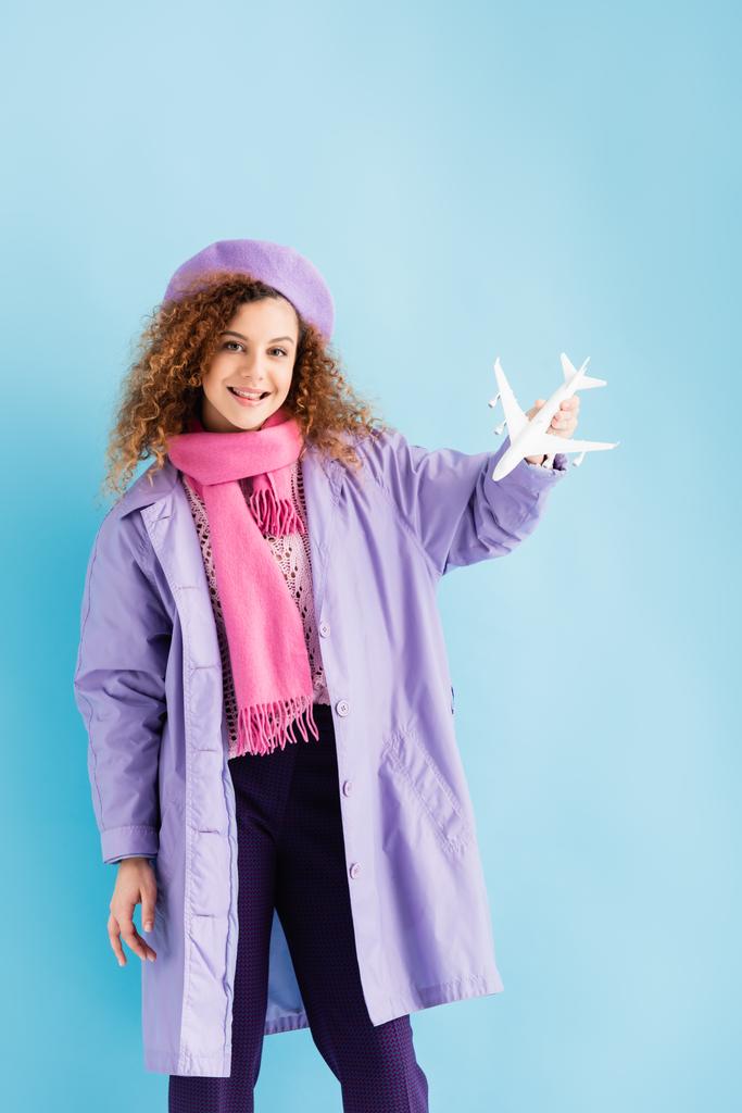 giovane donna positiva in berretto, sciarpa e cappotto in possesso di aereo giocattolo su blu - Foto, immagini