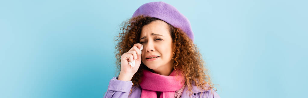 sconvolto giovane donna in cappotto invernale, berretto e rosa sciarpa lavorata a maglia che tiene il tessuto mentre piange sul blu, banner - Foto, immagini