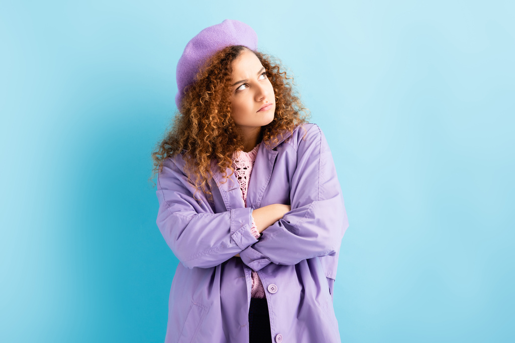 offended woman in beret looking away while standing with crossed arms on blue - Photo, Image