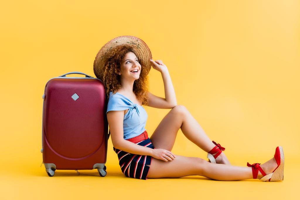 веселая и кудрявая женщина регулирует соломенную шляпу, смеясь и сидя рядом с чемоданом на желтом  - Фото, изображение