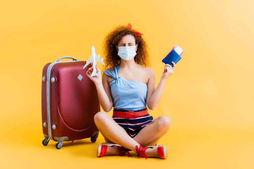 giovane donna riccia in maschera medica in possesso di passaporto e aereo giocattolo mentre seduto vicino alla valigia su giallo  - Foto, immagini