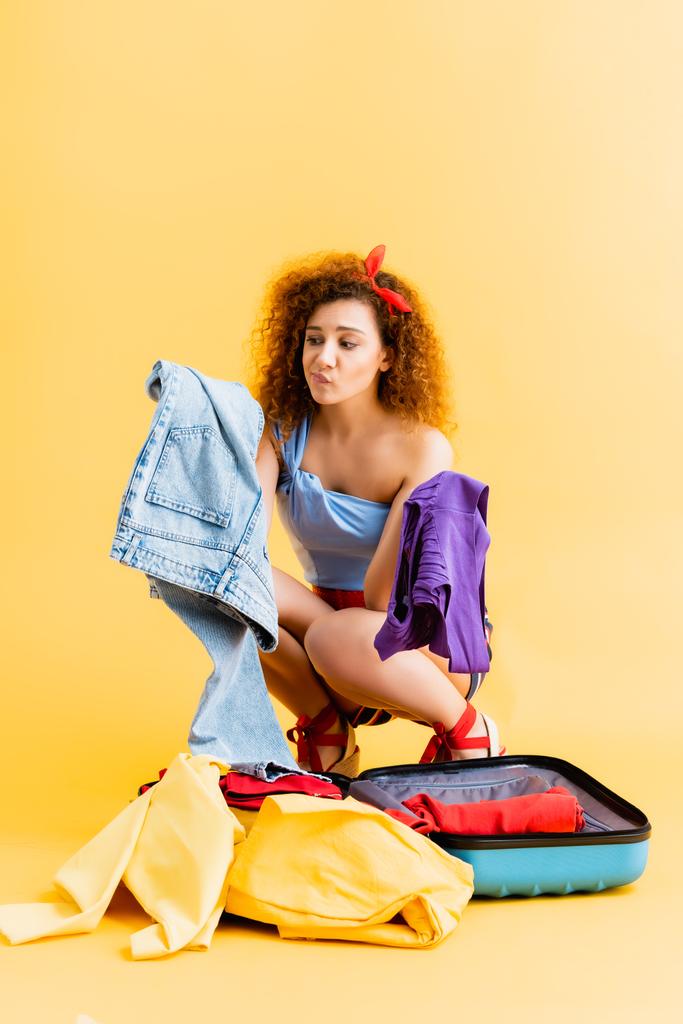 zmatená žena sedí u kufru a vybírá si mezi oblečením na žluté - Fotografie, Obrázek