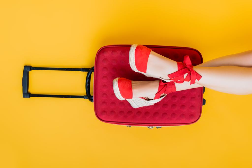 μερική άποψη της γυναίκας σε σφήνα σανδάλια κοντά σε κόκκινη βαλίτσα σε κίτρινο  - Φωτογραφία, εικόνα
