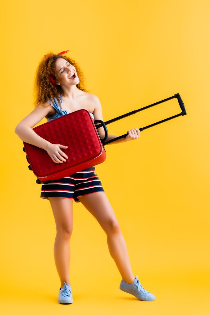 full length of happy woman in summer outfit and sneakers holding red suitcase and singing on yellow   - Photo, Image