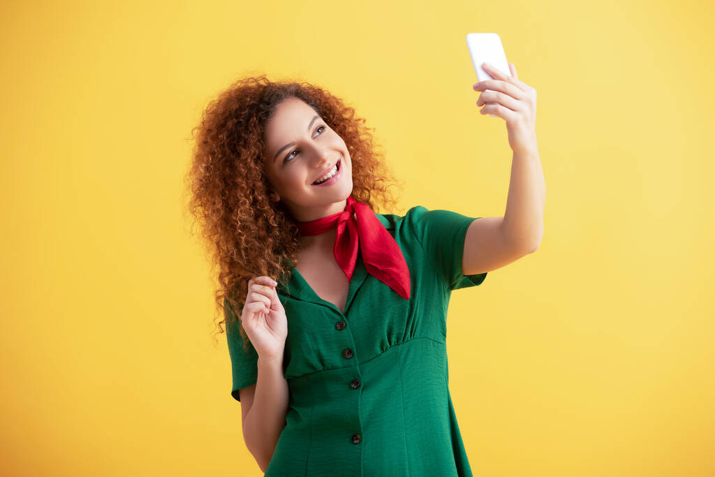 encaracolado jovem mulher em vestido verde sorrindo ao tomar selfie no amarelo  - Foto, Imagem