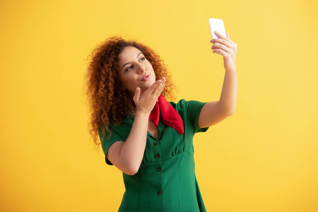 ricci giovane donna in abito verde invio bacio aria mentre prende selfie su giallo  - Foto, immagini