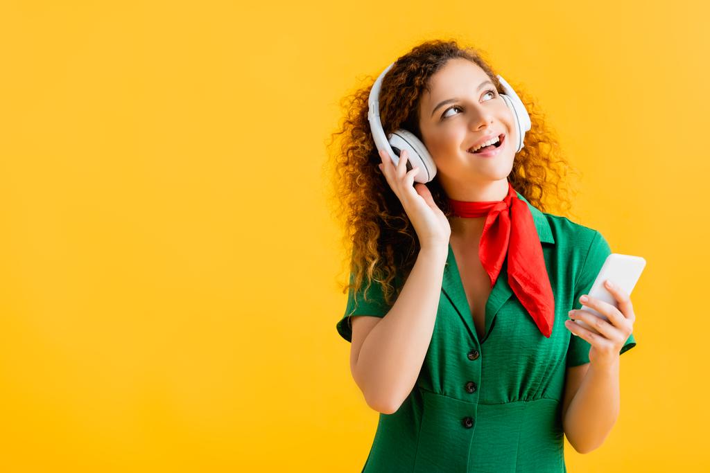 mujer alegre en auriculares inalámbricos sosteniendo teléfono inteligente aislado en amarillo  - Foto, imagen
