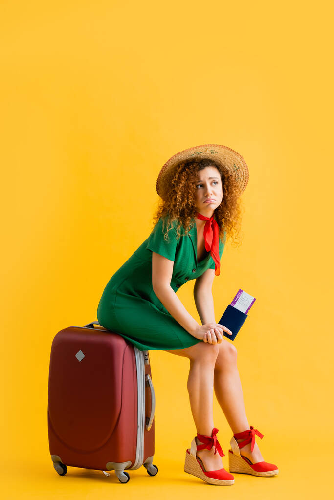 πλήρες μήκος της αναστατωμένης γυναίκας σε ψάθινο καπέλο κρατώντας διαβατήριο και κάθεται σε κόκκινες αποσκευές σε κίτρινο  - Φωτογραφία, εικόνα