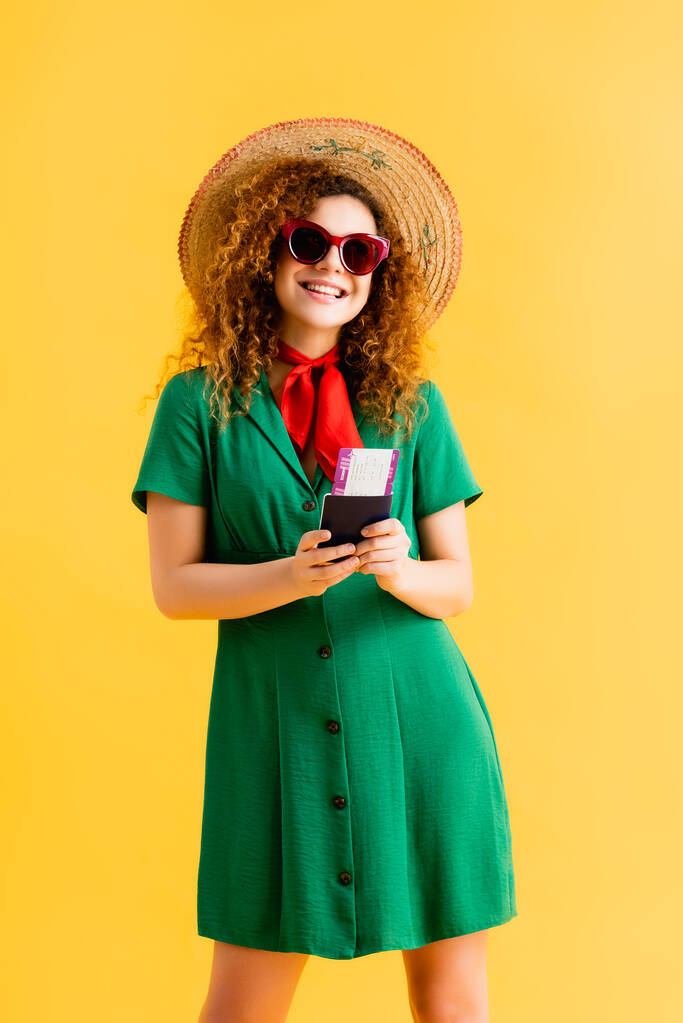 happy woman in straw hat, sunglasses and dress holding passport on yellow  - Photo, Image