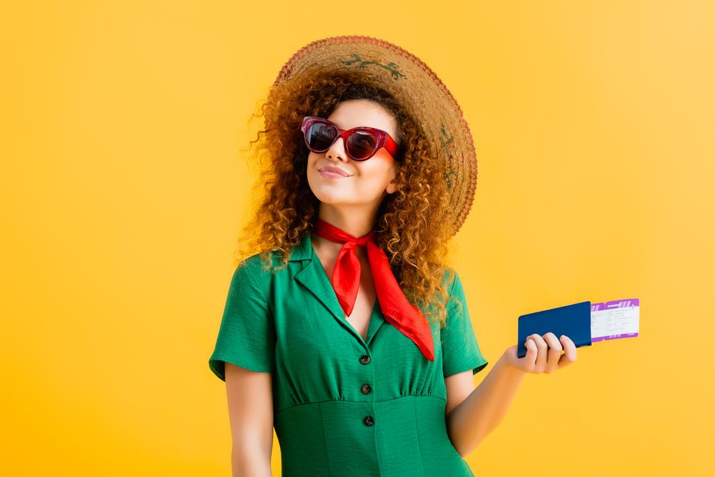 cheerful woman in straw hat, sunglasses and dress holding passport on yellow  - Photo, Image