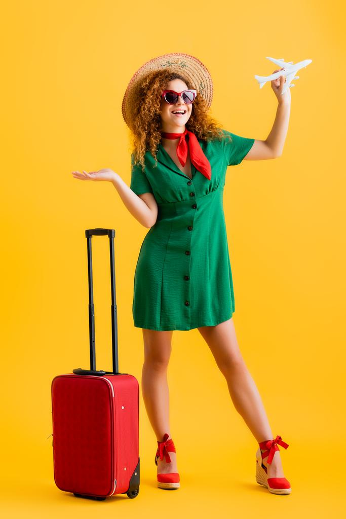in voller Länge glückliche Frau mit Strohhut, Sonnenbrille und Kleid, die Spielzeugflugzeug in der Nähe von Gepäck auf gelb hält  - Foto, Bild
