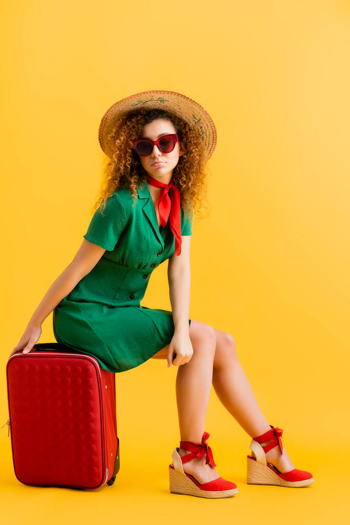 volledige lengte van trieste vrouw in stro hoed, zonnebril en jurk zitten op bagage op geel  - Foto, afbeelding