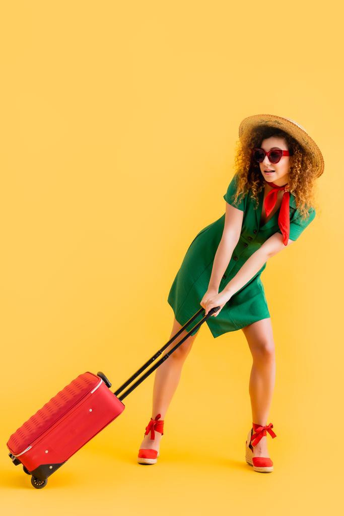 volledige lengte van de vrouw in stro hoed, zonnebril en jurk trekken bagage op geel  - Foto, afbeelding