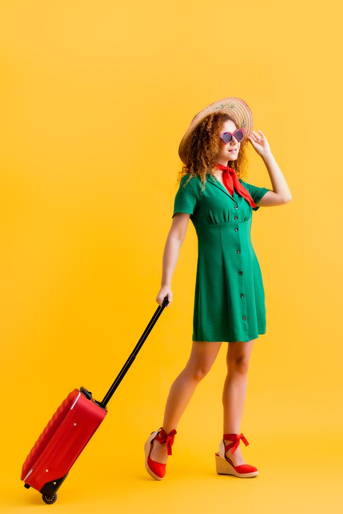 повна довжина кучерявої жінки в солом'яному капелюсі, сонцезахисних окулярах і сукні, що стоїть з багажем на жовтому
  - Фото, зображення