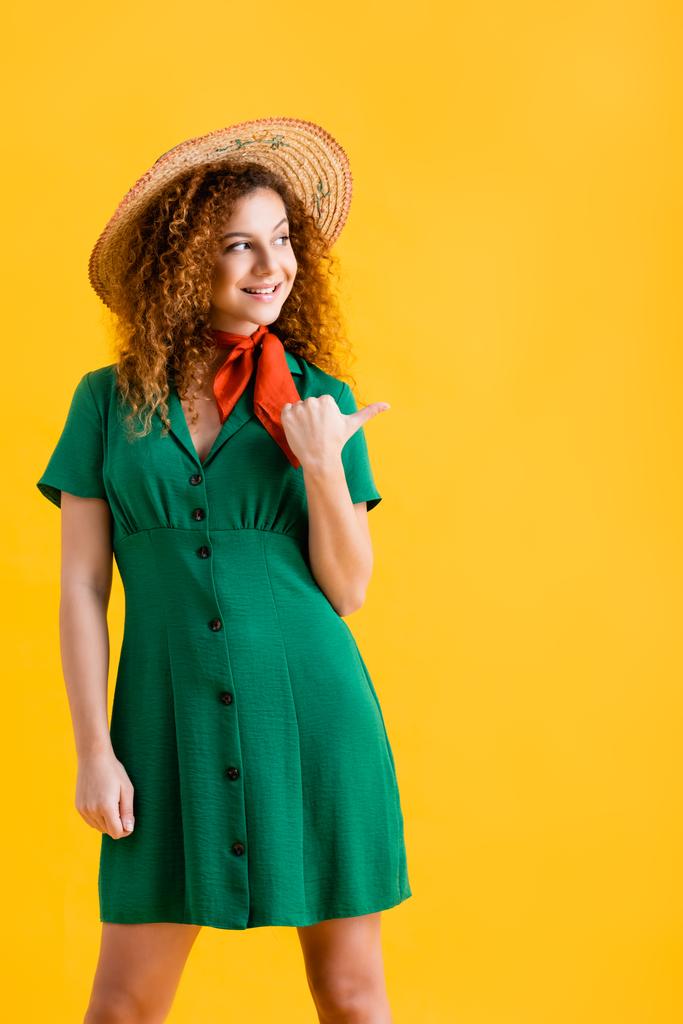 veselá mladá žena v slamáku a zelené šaty ukazuje palcem izolované na žluté  - Fotografie, Obrázek