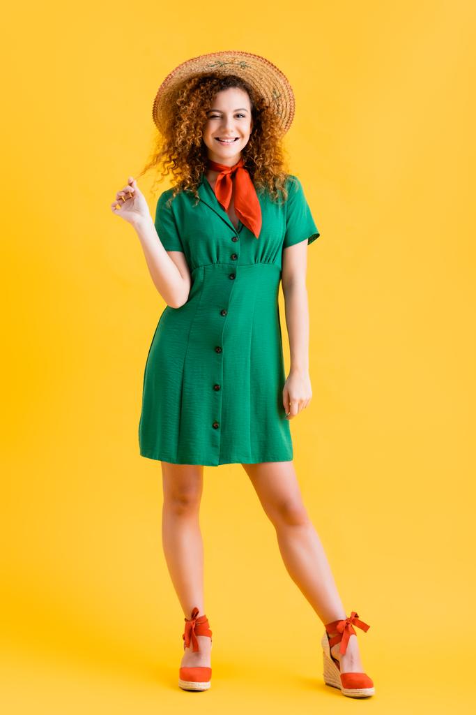 full length of cheerful young woman in straw hat and green dress standing on yellow  - Photo, Image