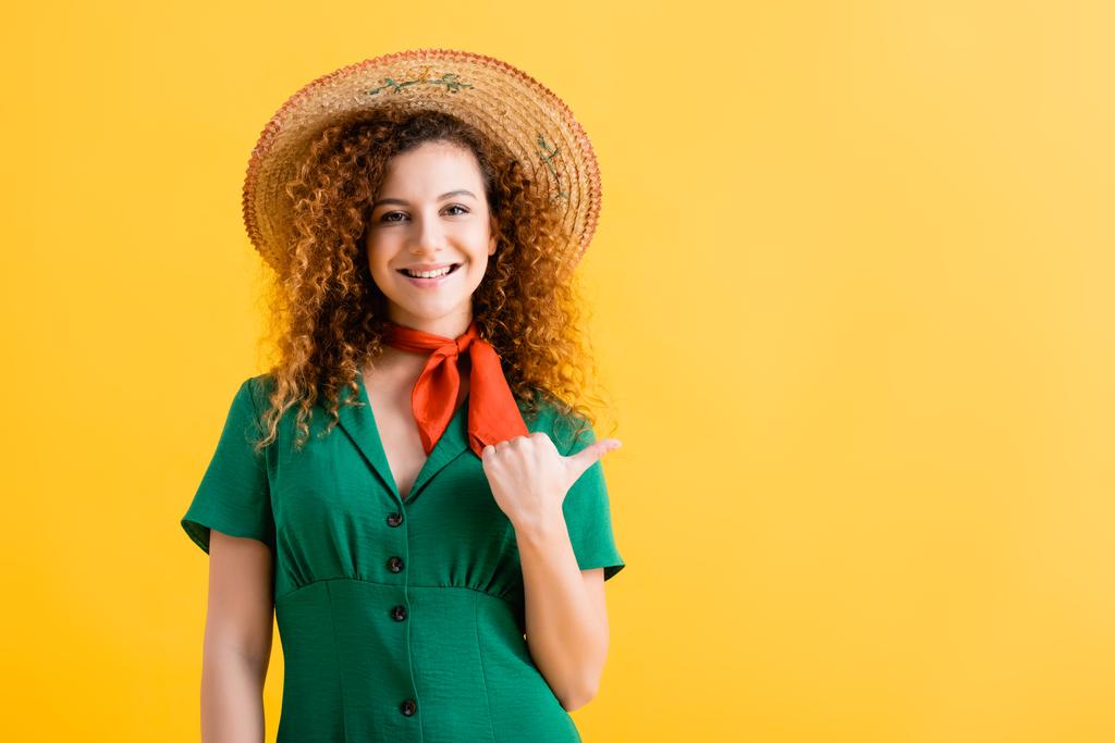 happy young woman in straw hat and green dress pointing with thumb isolated on yellow  - Photo, Image