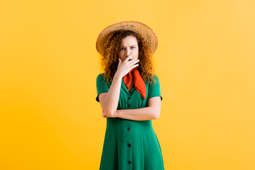 bang jonge vrouw in stro hoed en groene jurk bedekken mond op geel  - Foto, afbeelding