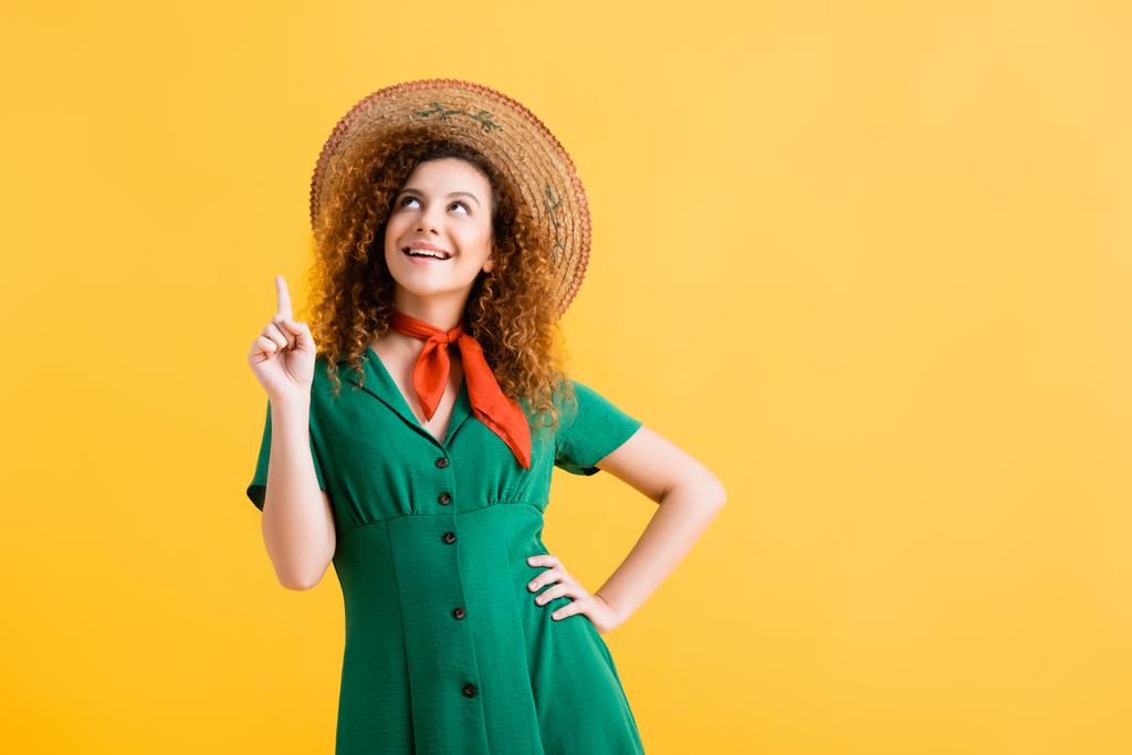 glückliche junge Frau mit Strohhut und grünem Kleid zeigt mit dem Finger und blickt auf gelb  - Foto, Bild