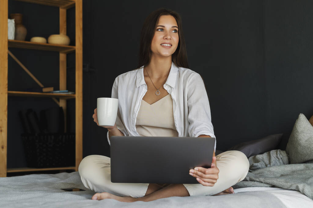 Millennial κορίτσι κάθεται στο κρεβάτι με λευκό πουκάμισο κρατώντας φλιτζάνι καφέ στο χέρι της. Όμορφη μελαχρινή γυναίκα χρησιμοποιεί φορητό υπολογιστή - Φωτογραφία, εικόνα