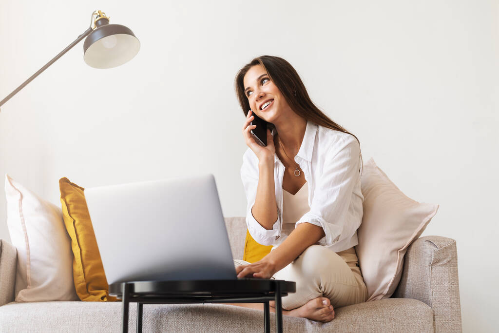 Atrakcyjna bizneswoman siedzi przy stole przed laptopem i rozmawia przez telefon komórkowy, negocjuje przez telefon.  - Zdjęcie, obraz