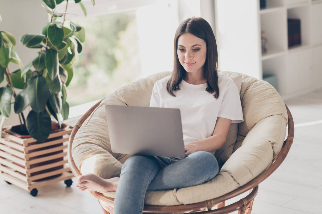 Fénykép okos lány ül fonott karosszék munka laptop visel fehér póló farmer a házban beltéri - Fotó, kép