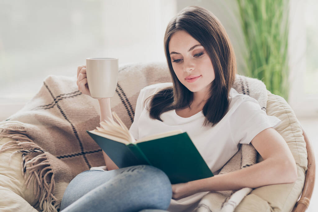 Φωτογραφία του εστιασμένου κορίτσι διαβάσει το βιβλίο για λυγαριά καρέκλα καρό κουβέρτα κρατήστε το φλιτζάνι τσάι στο σπίτι σε εσωτερικούς χώρους - Φωτογραφία, εικόνα