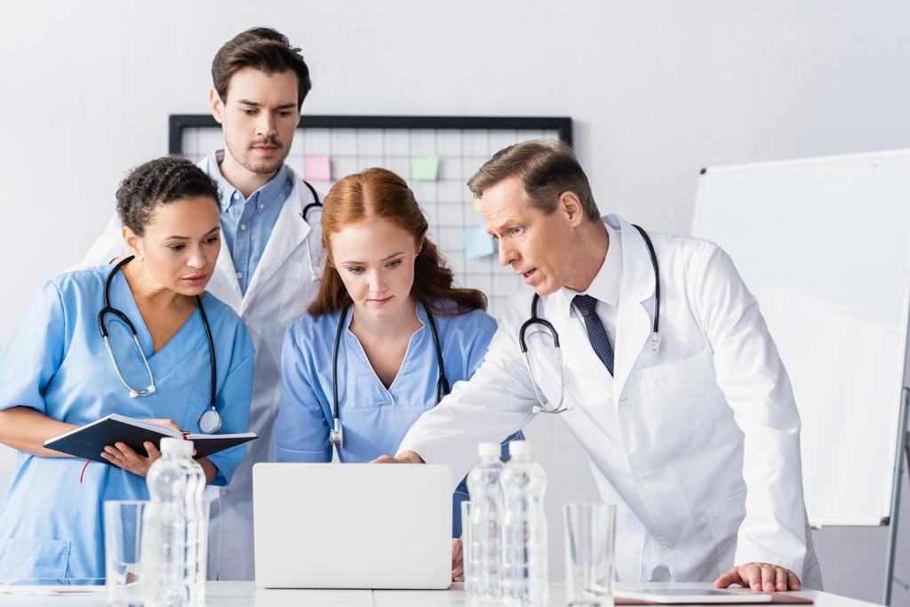 Médecins et infirmières multiethniques avec cahier regardant un ordinateur portable près de l'eau au premier plan flou  - Photo, image
