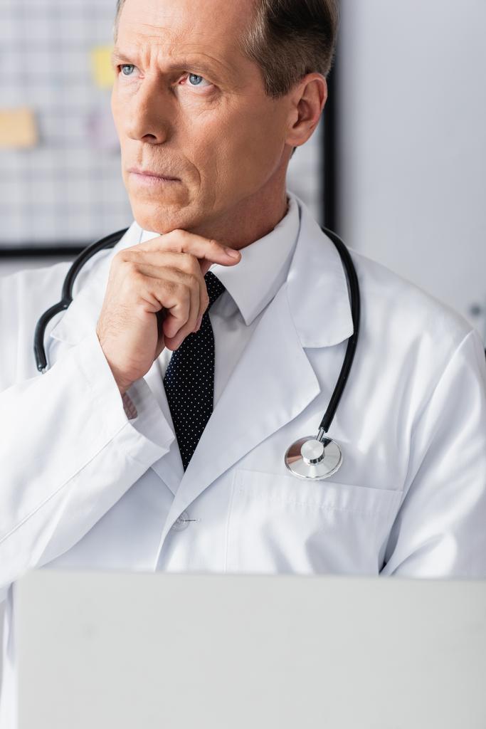 Przycięty widok przemyślanego lekarza trzymającego laptopa na niewyraźnym pierwszym planie w klinice  - Zdjęcie, obraz