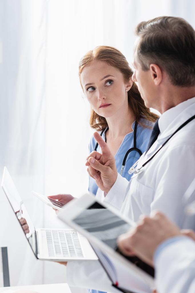 Doctor con portátil apuntando con el dedo cerca de la enfermera con tableta digital y colega en primer plano borrosa  - Foto, Imagen
