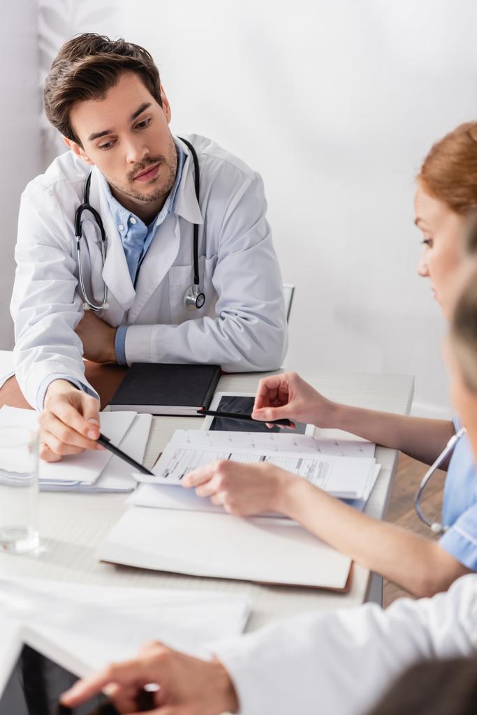 Lekarz wskazujący na papiery w pobliżu tabletu cyfrowego i pielęgniarki na niewyraźnym pierwszym planie  - Zdjęcie, obraz