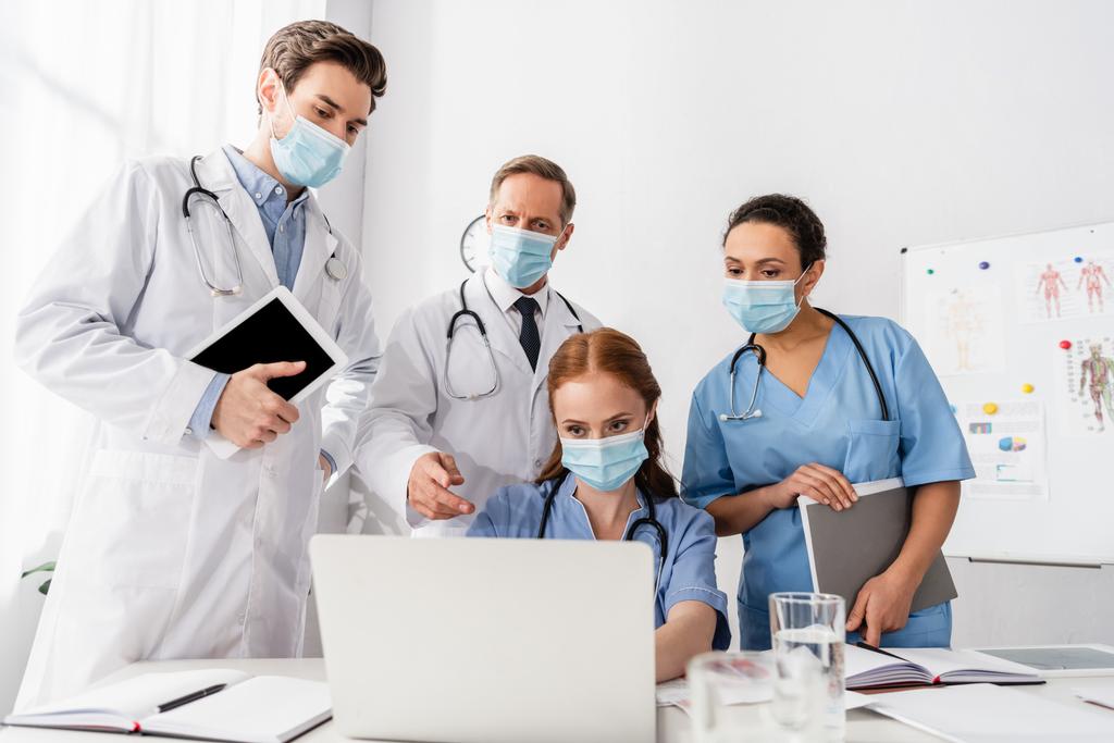 Tıp maskeli çok ırklı doktor ve hemşireler ön planda bulanık kağıtlar üzerinde çalışırken dizüstü bilgisayar kullanıyorlar.  - Fotoğraf, Görsel