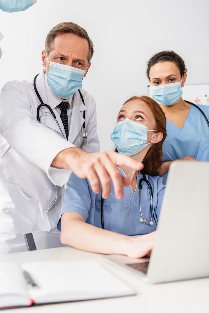 African american nurse standing near doctor pointing with finger at laptop near colleagues on blurred foreground - Photo, Image