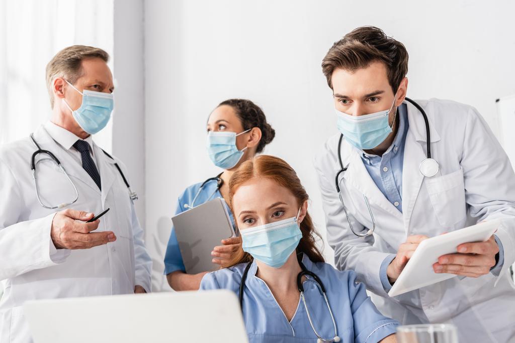 Sairaanhoitaja lääketieteellisessä naamiossa katselemassa kannettavaa tietokonetta lähellä monikulttuurisia kollegoita sairaalassa hämärtyneellä etualalla - Valokuva, kuva
