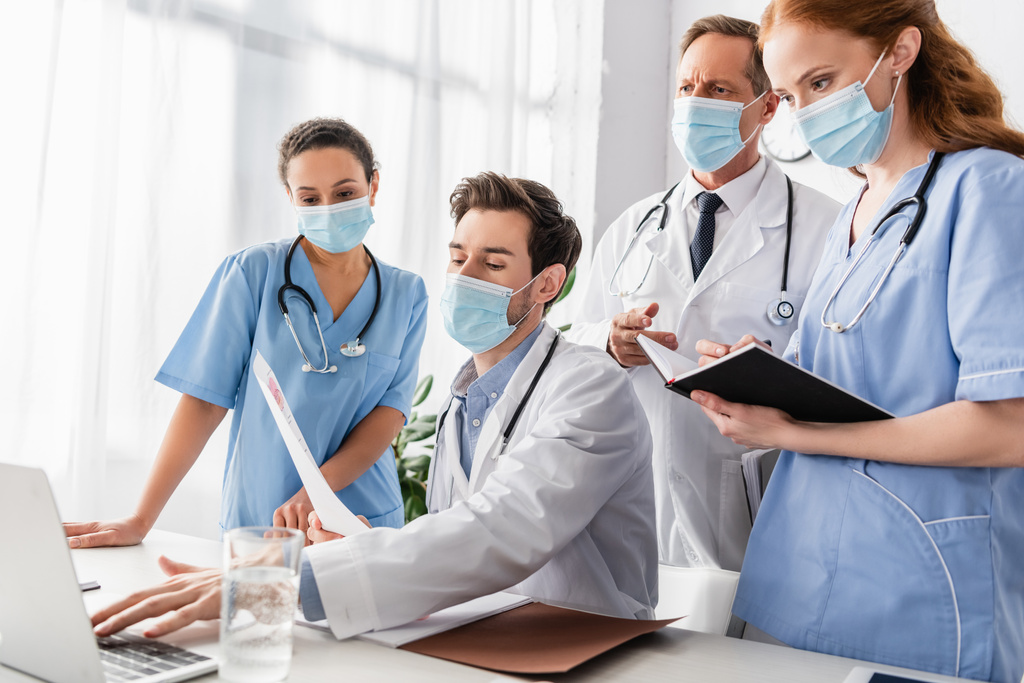 Colegas multiétnicos em máscaras médicas de pé perto médico digitando no laptop no local de trabalho no hospital - Foto, Imagem