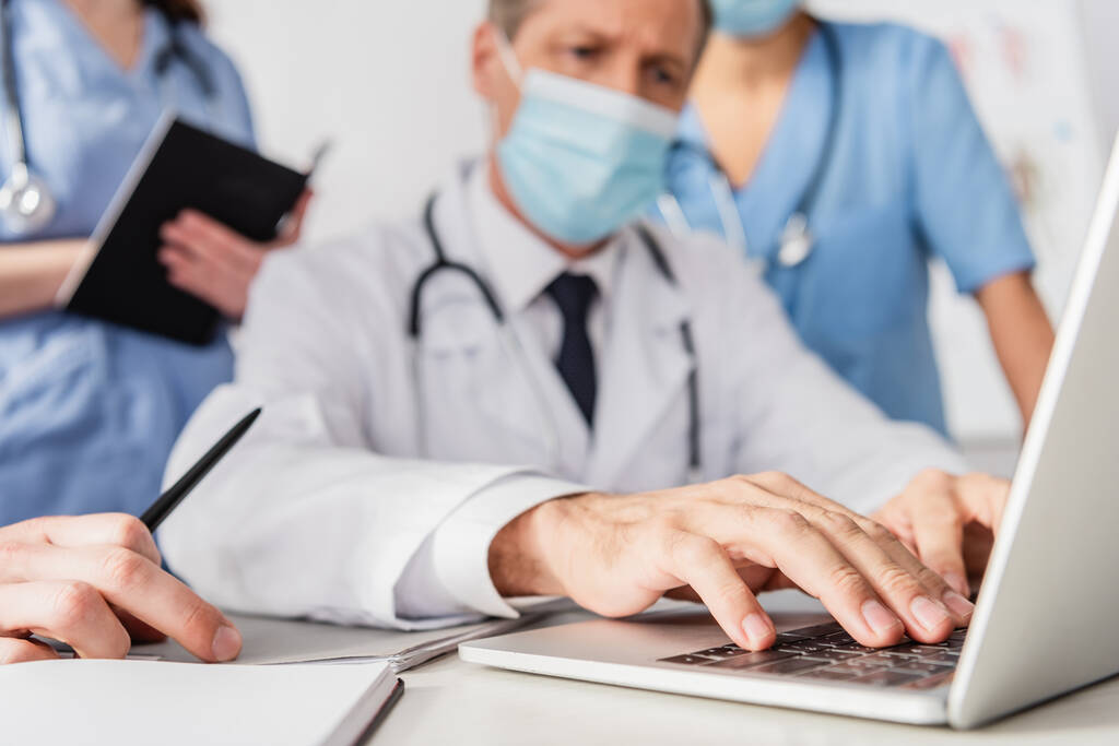 Крупный план врача в медицинской маске, печатающего на ноутбуке рядом с коллегами на размытом фоне - Фото, изображение