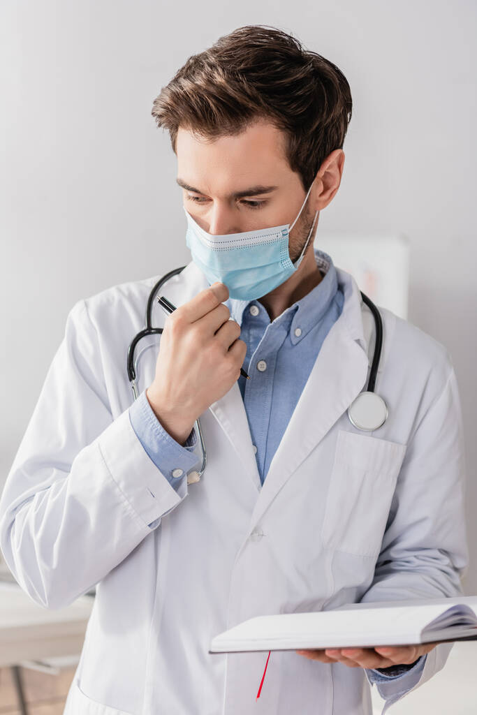背景がぼやけた状態でノートやペンを持ちながら医師が医療用マスクに触れる様子 - 写真・画像