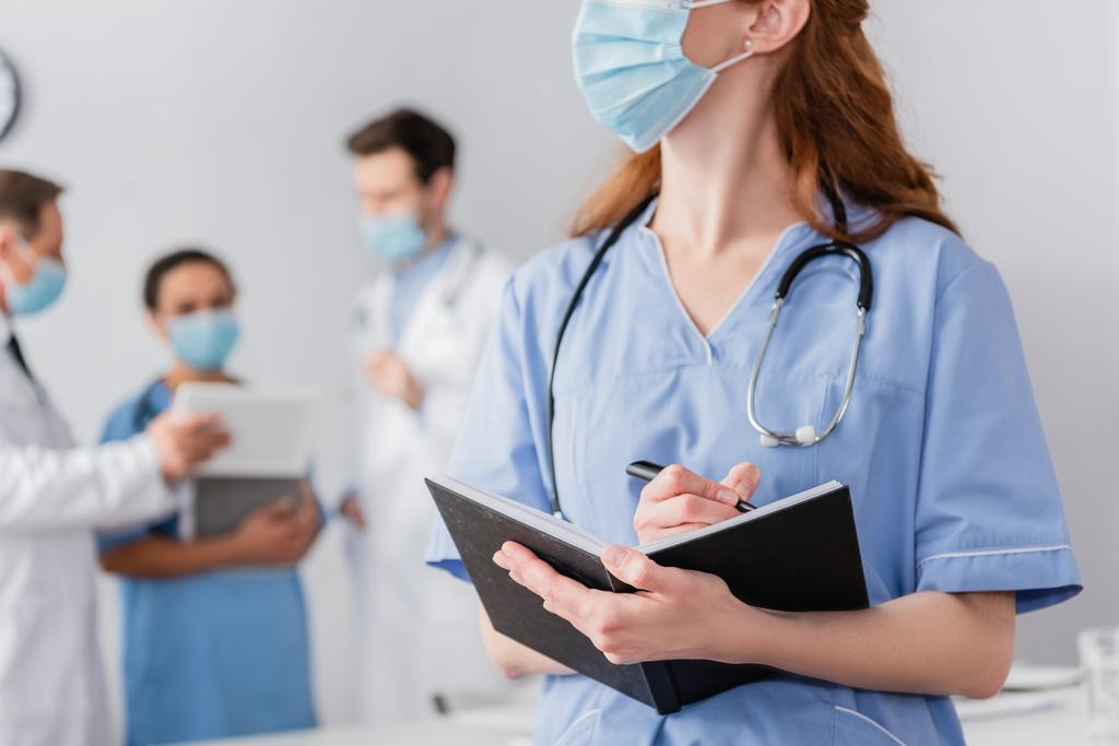 Руда медсестра в медичній масці пише в блокноті з розмитими багатоетнічними колегами, що працюють на фоні
 - Фото, зображення