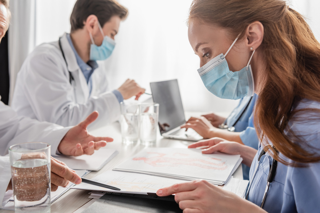 Rothaarige Krankenschwester schaut auf Papiermappe mit Bildern, während sie in der Nähe des Arztes sitzt und mit Stift auf Kollegen auf verschwommenem Hintergrund zeigt - Foto, Bild