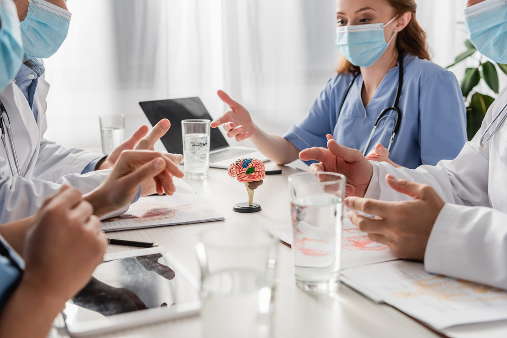 Médicos e enfermeiros em máscaras médicas conversando enquanto sentados no local de trabalho com dispositivos, fotos e copos de água em primeiro plano turvo - Foto, Imagem